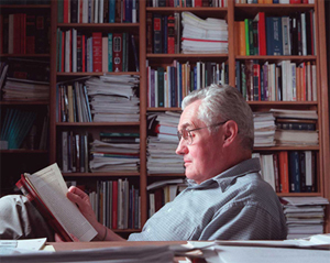 Barton, seated profile to the camera, reads a book in front a large, filled bookshelf.