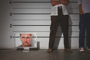 Photo-illustration of men in a prison line-up; a computer monitor depicting a man's face is included in the line-up.