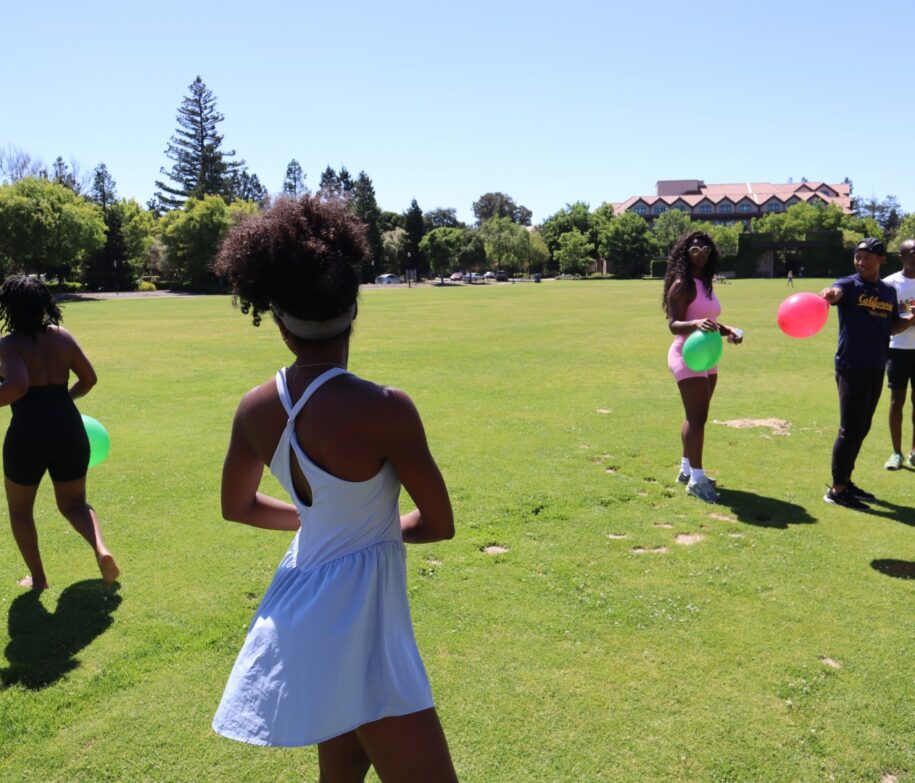 Image shows four students in a water balloon fight on a large green field.