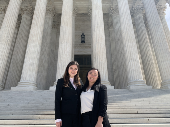 Julia on the left and Victoria on the right stand together wearing professional attire and smiling with the tall marble pillars and steps of the Supreme Court behind them.