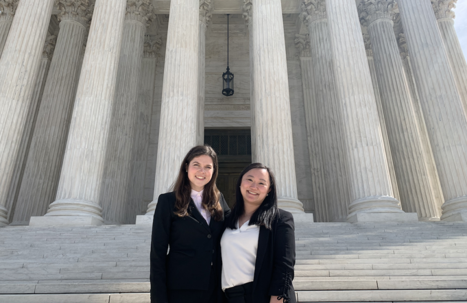 Julia on the left and Victoria on the right stand together wearing professional attire and smiling with the tall marble pillars and steps of the Supreme Court behind them.
