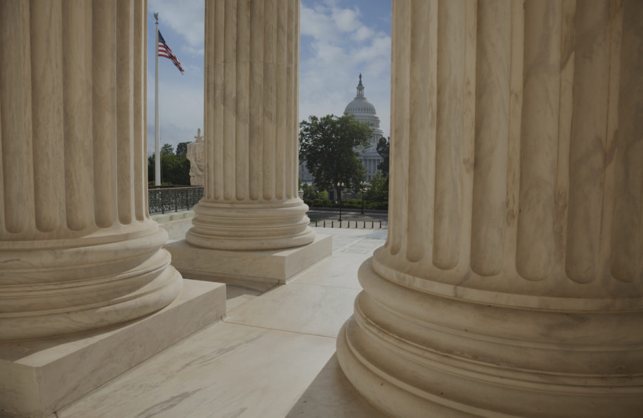 Close up of the columns of the Supreme Court building with an American flag and the US Capitol in the background