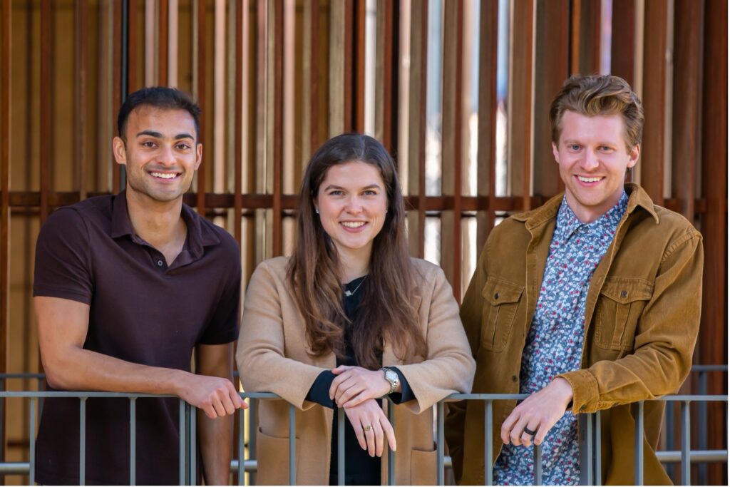 From left to right: Seraj, Julia, and Tanner are standing in front of a wooden wall, smiling while leaning their arms on a metal balcony