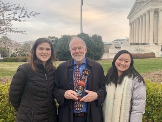 From left to right, Julia, Phil, and Victoria are wearing warm winter coats and smiling in front of green bushes and the lawn on the rightside of the Supreme Court building. Phil is holding the “Bad Spaniels” dog toy.