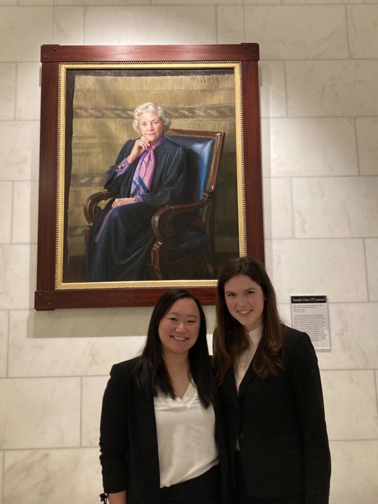 Victoria on the left and Julia on the right are wearing professional attire, smiling together, and standing in front of a wood-framed portraitof Justice Sandra Day O’Connor sitting in a chair