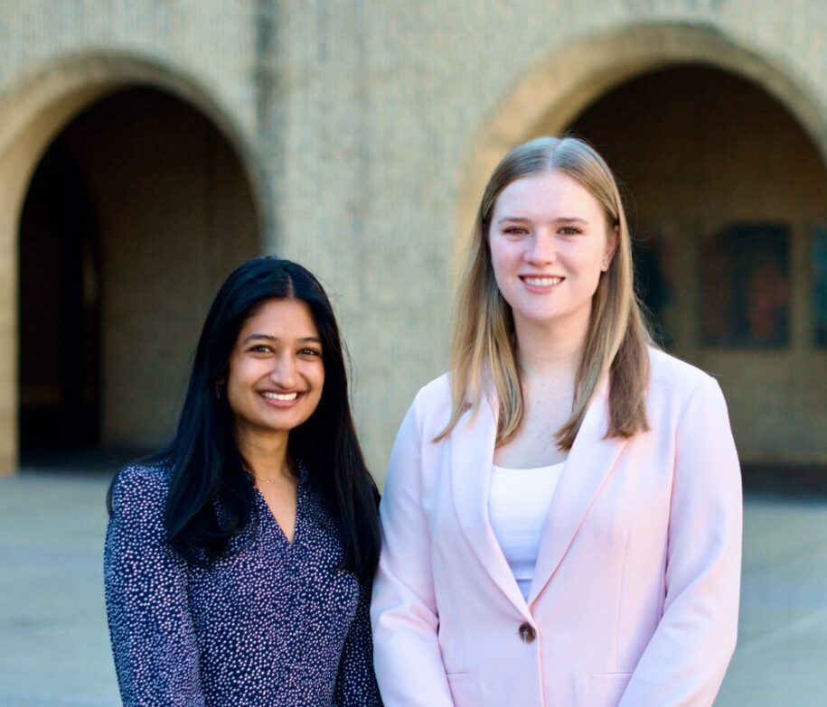 SLS students Kiran and Victoria stand side by side, facing the camera. In the background is the blurry image of two arches on the SLS Crown building.
