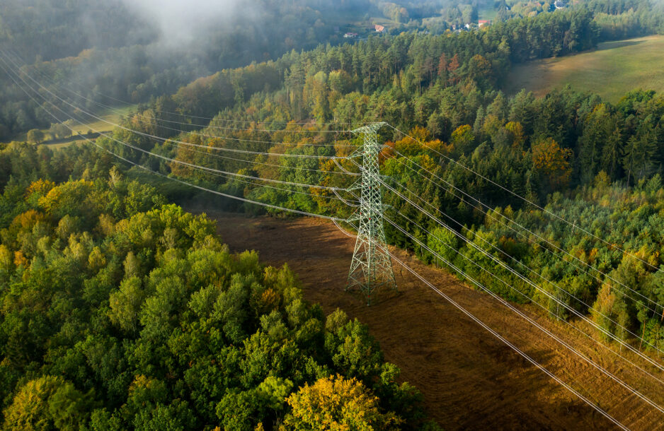 Aerial view of the high voltage power lines and high voltage electric transmission tower on the terrain path surrounded by trees.
