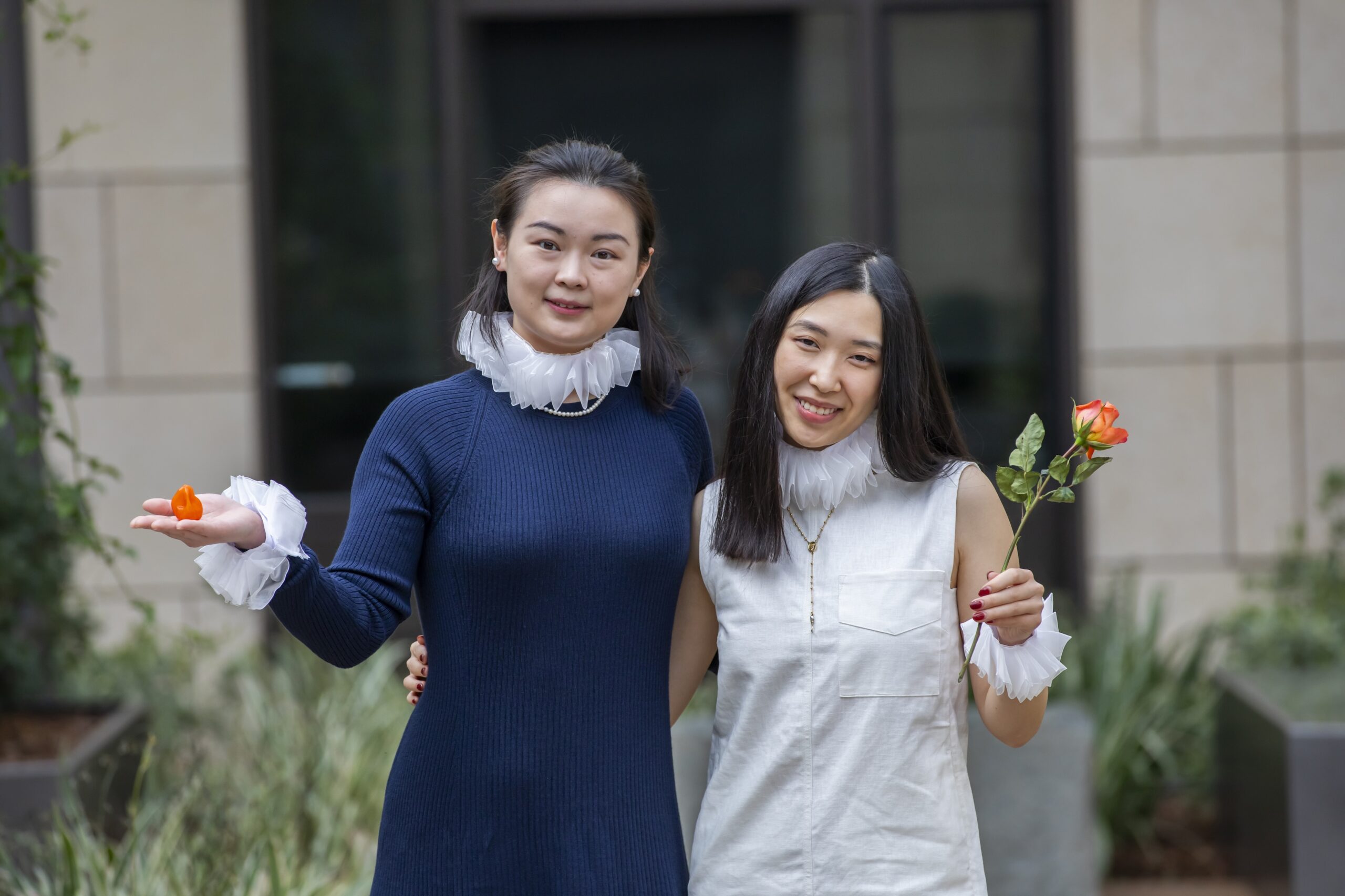 Isabella in a blue dress standing to the left of Selina in a white dress, each wearing white Elizabethan collars and wrist cuffs and holding a pepper and rose in their hands