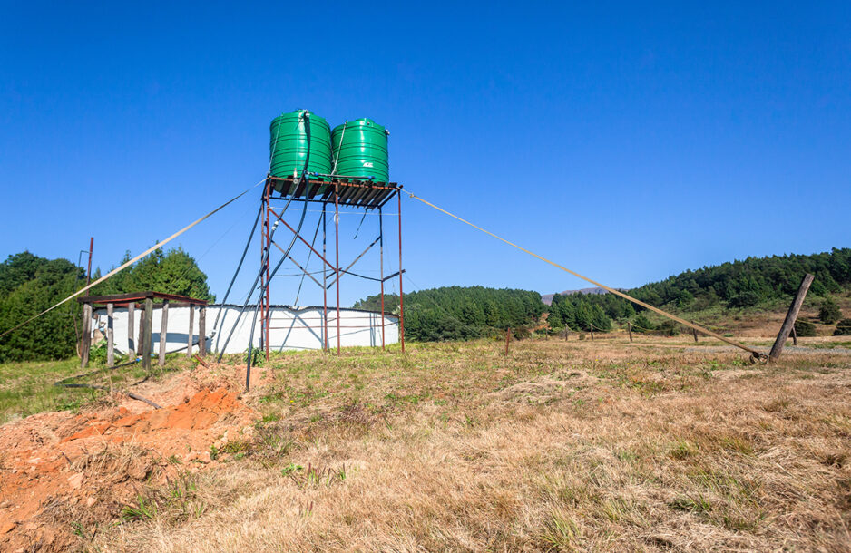 Two large green colored water tanks on tall steel stand in open field in blue sky landscape.