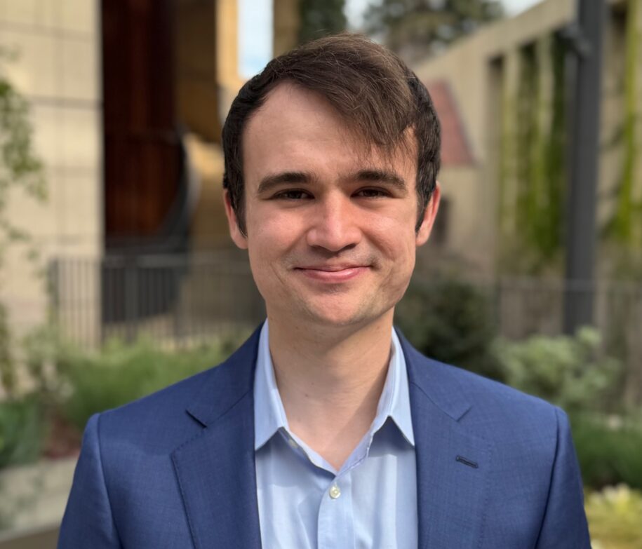 August faces the camera wearing a light blue shirt and dark blue jacket