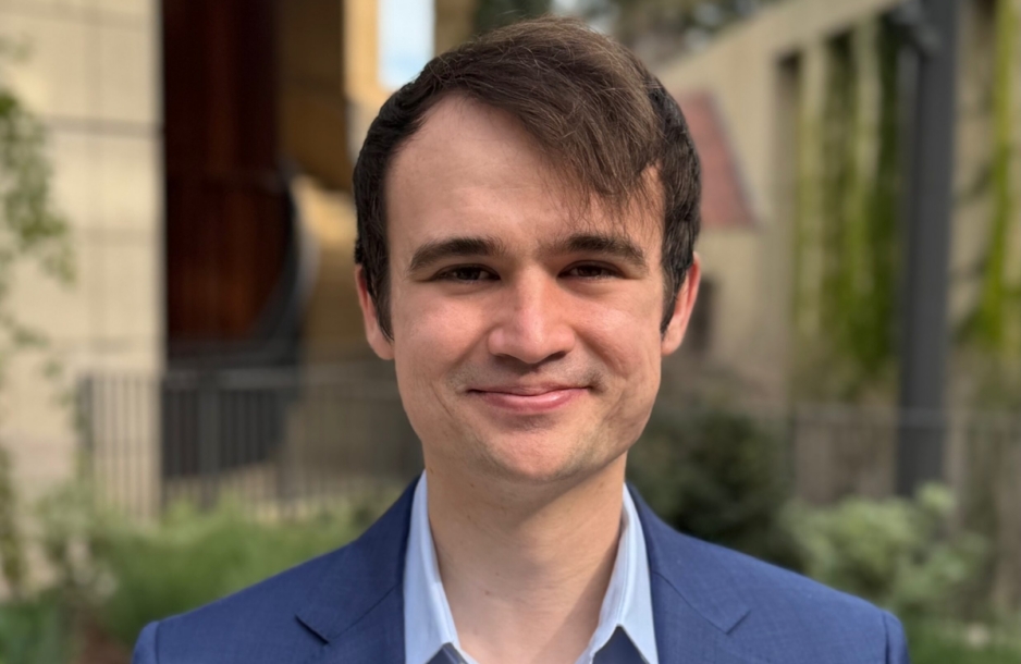 August faces the camera wearing a light blue shirt and dark blue jacket